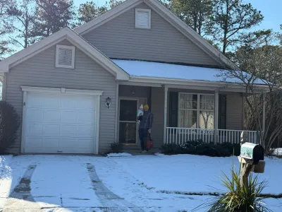 A snow-covered ranch home exterior with gray siding, white garage door, and a person holding a child on the porch.
