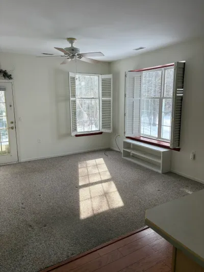 Empty living room with worn carpet, bay window, plantation shutters, and a built-in window seat.