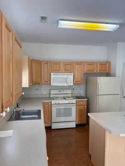 Dated galley kitchen with light oak cabinets, white appliances, and laminate countertops before renovation.