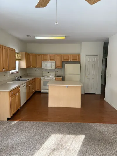 Dated kitchen with light oak cabinets, white appliances, and a small island, transitioning into a carpeted living area.