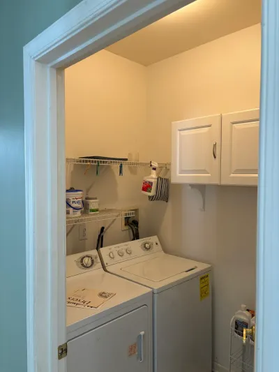 A small laundry closet with a washer and dryer, wire shelving, and a white wall cabinet before renovation.