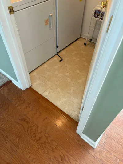 A laundry closet with worn vinyl flooring and a white dryer, seen through a doorway from a hardwood-floored hallway.