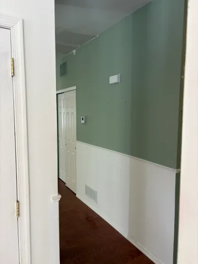 A narrow hallway with sage green upper walls, white lower walls, and dark wood flooring, seen through a doorway.