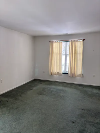 Empty room with worn green carpet, off-white walls, and a window dressed in cream curtains.