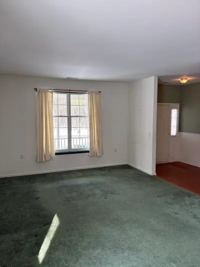Empty living room with green carpet and curtained window, opening into a hallway with wood flooring.