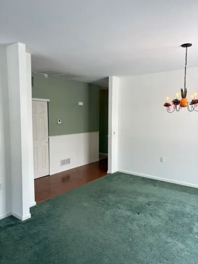 Empty room with green carpet, sage and white walls, and a decorated wrought iron chandelier before renovation.