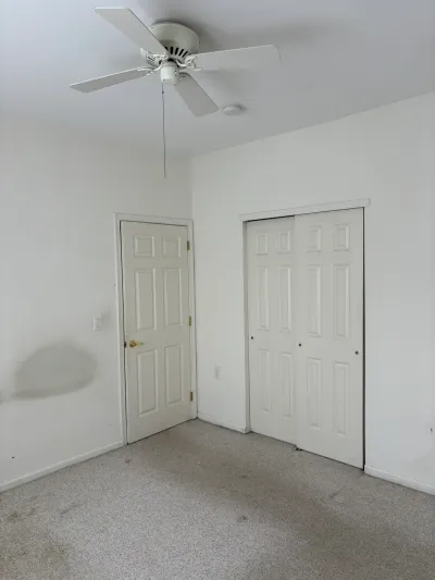 Empty bedroom with beige carpet, white closet doors, and a ceiling fan, shown in pre-renovation condition.