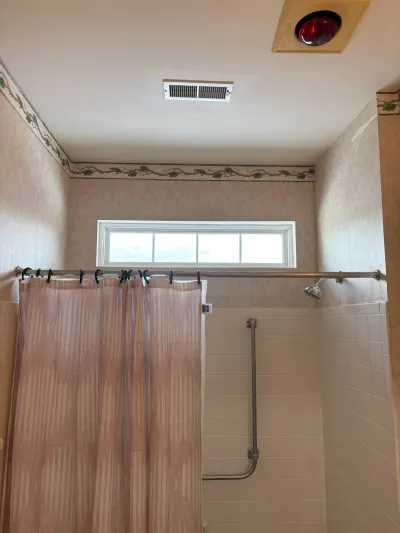 An outdated shower stall with beige tile, floral wallpaper border, and exposed chrome plumbing before renovation.