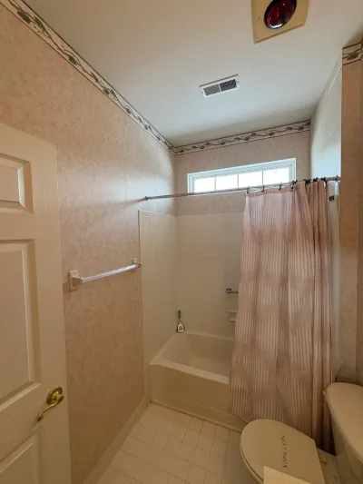 Dated bathroom with wallpaper, tiled tub-shower alcove, and a toilet still wrapped for sanitation before renovation.