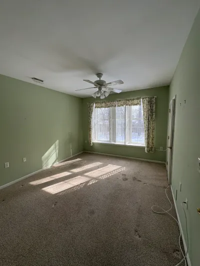 Empty bedroom with green walls, worn carpet, and a window fitted with plantation shutters before renovation.