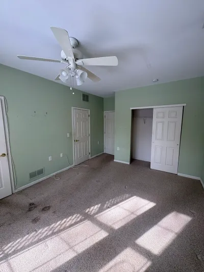 Empty bedroom with sage green walls, worn gray carpet, and a ceiling fan, before renovation.