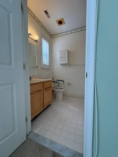 Dated bathroom with floral wallpaper, tile floor, wood vanity, and a toilet fitted with a raised safety seat.