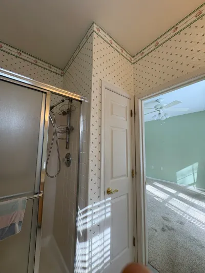 Dated bathroom with floral wallpaper and glass shower stall, doorway open to a carpeted room beyond.
