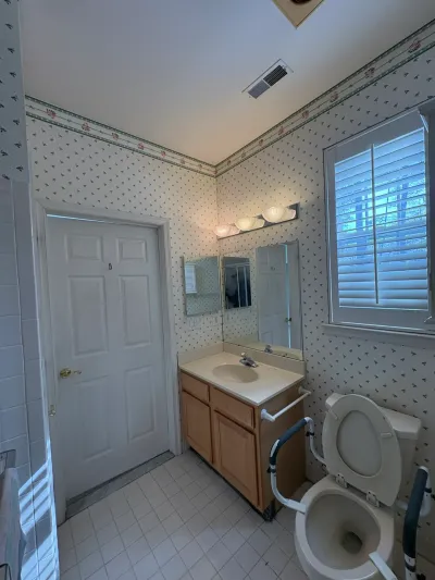 Dated bathroom with floral wallpaper, wood vanity, and a toilet fitted with safety grab bars.