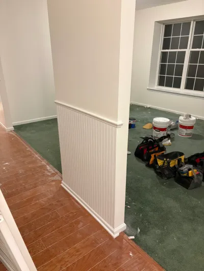 A basement under renovation with new white wainscoting, paint buckets, and tool bags on green carpet.