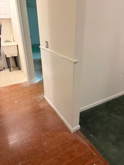 Basement hallway under renovation with new beadboard wainscoting and a floor transition from wood-look flooring to carpet.