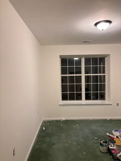 A partially painted bedroom at night with green carpet, paint supplies on the floor, and a lit ceiling fixture.
