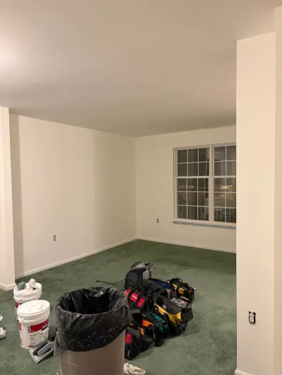 A basement room mid-renovation with fresh paint, old green carpet, and contractor tools and buckets on the floor.