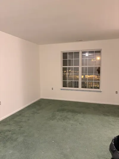 Empty room with worn green carpet and a large window, photographed before renovation work begins.