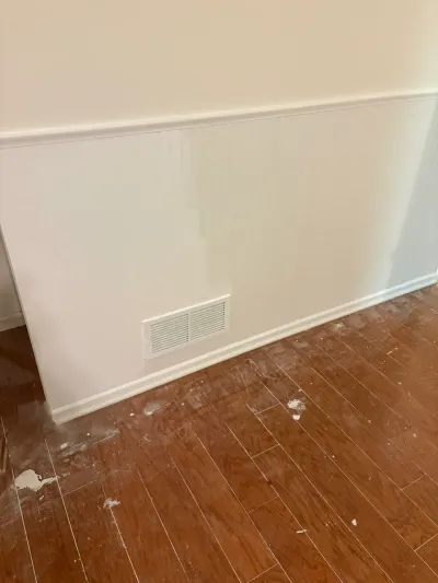 Wall corner with chair rail molding and a floor vent, wood floor below marked with paint splatter.