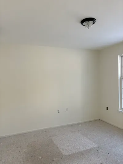 Empty bedroom before renovation with worn carpet, cream walls, and a black ceiling light fixture.