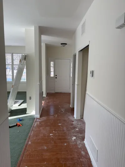 A hallway mid-renovation with new wood flooring, wainscoting, and a ladder visible in an adjoining carpeted room.