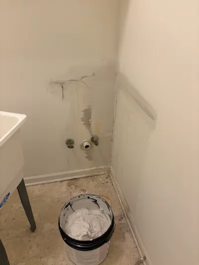 Laundry room wall with water stains, exposed plumbing valves, and a bucket of drywall compound mid-repair.