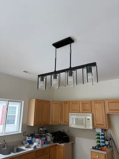 Kitchen mid-renovation with a new black glass-shade chandelier installed above oak cabinets and cluttered counters.