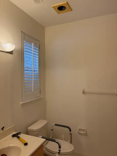 A bathroom mid-renovation with an unfinished ceiling light, a toilet safety rail, and tools on the counter.