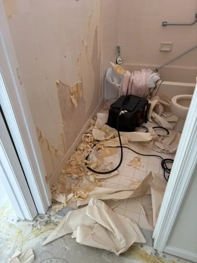 Bathroom mid-renovation with torn wallpaper strips scattered across the tile floor and pink tile tub surround.