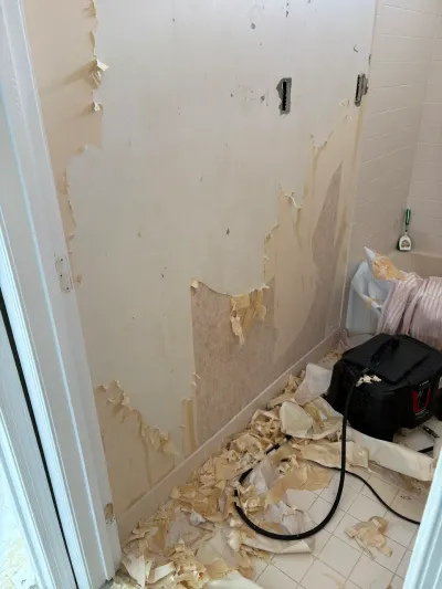 Bathroom wall mid-renovation with peeling wallpaper and debris scattered on the tile floor.