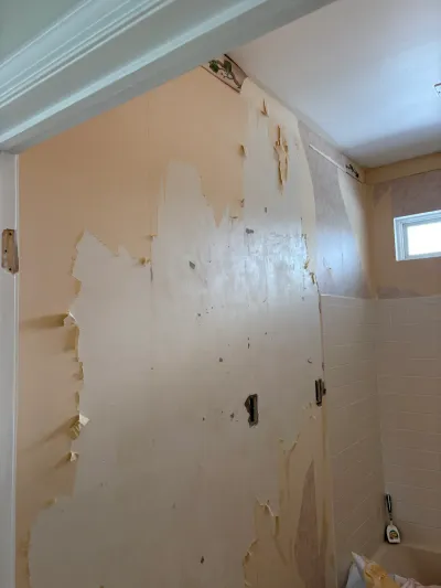 Bathroom wall with wallpaper being stripped away, revealing peach paint and bare drywall beside a tiled tub surround.