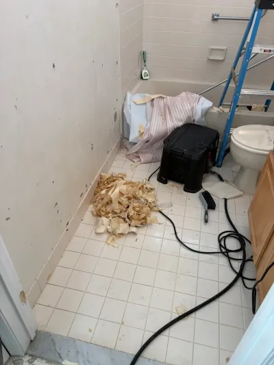 Bathroom mid-renovation with wallpaper stripped from walls and debris piled on the tile floor.