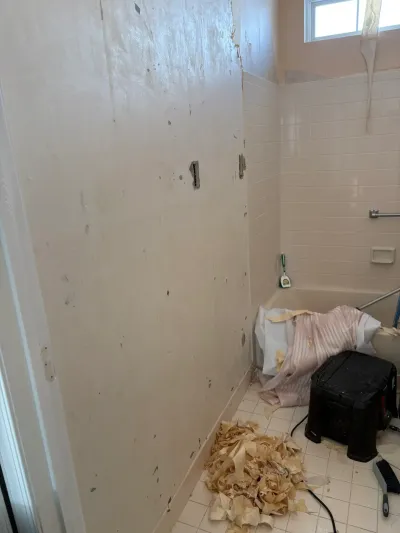 A bathroom mid-renovation with wallpaper being stripped from the wall and debris scattered on the tile floor near the tub.