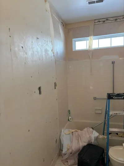 Bathroom mid-renovation with wallpaper being removed above a tiled tub surround and a ladder inside the tub.