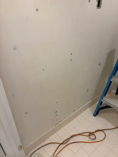 A bathroom wall mid-renovation with scraped drywall, a stepladder, and extension cords on tile flooring.