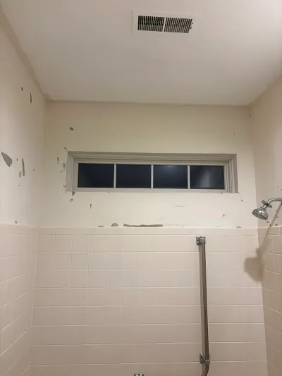 Bathroom shower before renovation, showing patchy tile, worn paint, and a window built into the shower wall.