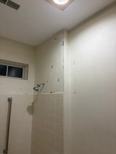 Bathroom shower wall with peeling paint above cream tile, a chrome showerhead, and a small window.