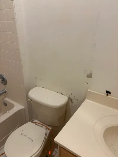 A dated beige bathroom before renovation, showing peeling wall paint and a toilet with a broken seat label.