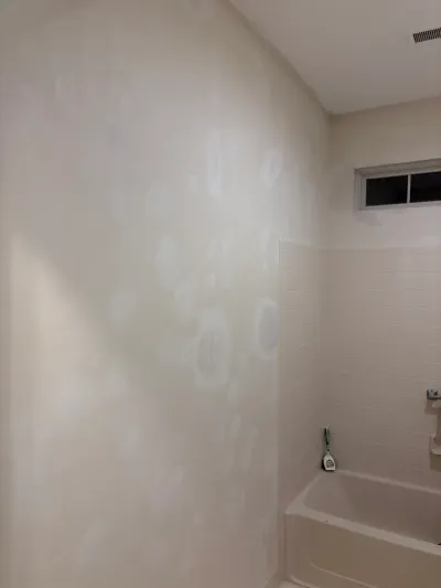 Bathroom wall with drywall patches next to a beige subway tile tub surround and window.