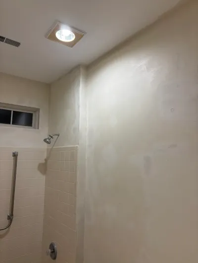 Shower wall mid-renovation with tiled lower half and patched drywall above, showerhead and grab bar visible.