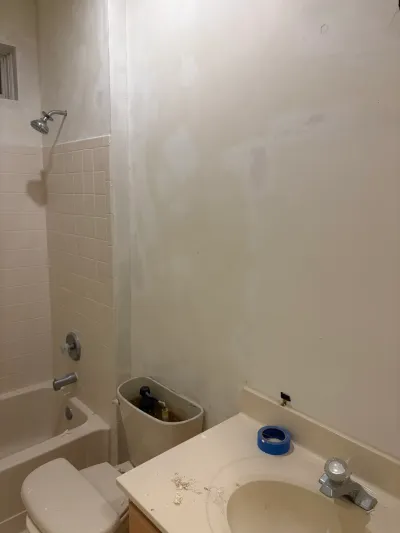 A bathroom mid-renovation with a disconnected toilet, patched drywall, and a tub/shower surround with beige tile.