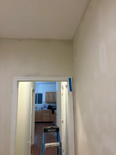 Hallway with beige walls under repair, leading through a doorway into a kitchen with wood cabinets.