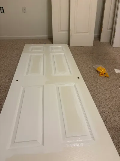 A freshly painted six-panel door lies on carpet in a basement, with more doors leaning against the wall behind it.