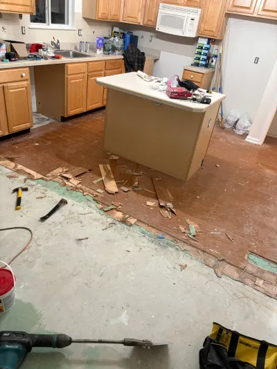 A kitchen mid-renovation with flooring torn up to bare concrete and demolition tools scattered on the floor.