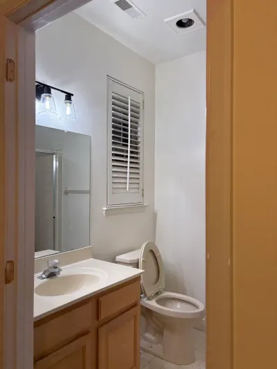 A small dated bathroom with an oak vanity, black sconce lighting, and a shuttered window above the toilet.