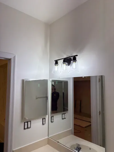 A black three-bulb vanity light glows above corner-mounted mirrors in a bathroom mid-renovation.