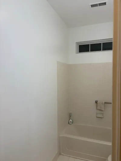 A dated bathroom with a beige tiled tub surround and a small window above it, before renovation.