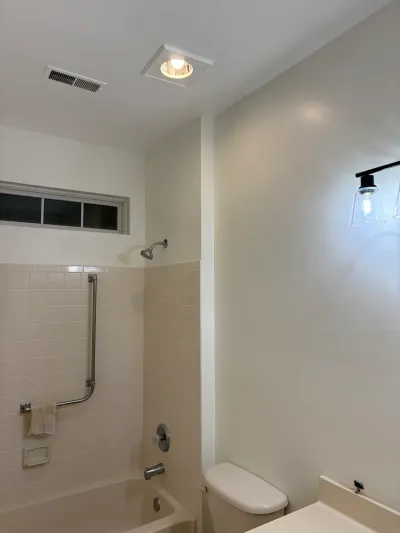 A dated beige-tiled bathtub and toilet sit beside a wall with a newly installed modern black sconce light.