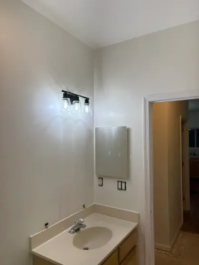A bathroom mid-renovation with a new vanity light, unmounted mirror, and exposed sink plumbing.